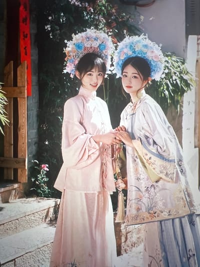 Two young East Asian women are standing outdoors in what appears to be a traditional Chinese courtyard, likely in the city of Liandu District, China. The woman on the left is wearing a pale pink, long-sleeved traditional garment over a light-colored skirt. She has a floral headdress adorned with pink and blue flowers. The woman on the right is wearing a similarly styled outfit, with a more intricate, patterned robe in light purple and blue tones, also featuring a floral headdress in blue and pink. Her robe has detailed embroidery of flowers and birds. The women are holding hands and appear to be interacting. They are positioned on stone steps in front of a weathered brick wall and lush greenery. To the left, a wooden structure with red banners hangs. The scene evokes a sense of cultural heritage and possibly a ceremonial occasion.