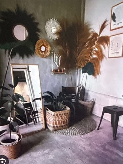 The image depicts a bohemian-style interior scene, likely a living room or a styled corner of a home. The backdrop is a textured gray wall. On the wall, there are three decorative round items: a large dark green, spiky circular piece with a central oval mirror, a smaller white fluffy circular piece, and a tan woven circular piece with a white center. To the right of these, a macrame hanging decoration with two small shelves is suspended. Further to the right on the wall are two framed art prints with minimalist line drawings.

In the foreground, a tall floor mirror with a thin frame leans against the wall. In front of the mirror are several potted plants. One large plant with dark green leaves is in the foreground left, in a round, woven basket planter. To the right, another plant in a similar woven basket planter is partially visible. A woven circular rug is placed on the floor in front of the plants and the mirror.

Behind the plants, a dark wooden chair is positioned. Large dried palm fronds are arranged in a vase or holder, creating a dramatic, feathery element. Another basket is visible behind the chair. A dark metal stool with three legs stands to the right of the scene. The flooring appears to be concrete with a slightly mottled purple and gray finish.

The image is a still photograph, taken with a mobile device, evidenced by the camera icon in the bottom left corner. There are no people visible in the scene, and no actions are being performed. The environment suggests a curated, indoor decorative setting.

The location in the city of Babahjurong, Indonesia, is not determinable from the visual information presented in the image.
