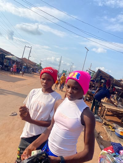 This image depicts an outdoor market scene in Amaseri, Nigeria, on a sunny day. In the foreground, two young women stand on an unpaved, reddish-brown dirt road, facing the camera. The subject on the left wears a red beanie with "AMIRI" in white text, a white short-sleeved t-shirt with a faded graphic (resembling a designer logo), and dark pants/shorts. Her left hand is partially closed, holding a small, indistinct object. The subject on the right wears a pink beanie with colorful letters spelling "HAPPY" (partially visible), a white ribbed sleeveless turtleneck top, and blue patterned shorts. Her left hand holds several green Nigerian Naira notes (specifically 100 Naira denominations are visible) and a black plastic bag containing items.
In the midground and background, the street extends, lined with various rudimentary market stalls constructed from wood, corrugated metal, and tarps. These stalls display merchandise such as clothing and other household goods. Several individuals are visible walking or tending to the stalls. On the far right, a man in a dark blue shirt and green striped pants bends over a wooden table at a stall. Utility poles with numerous overhead power lines crisscross the sky. A tall, lattice-structured communication tower is prominent on the right side of the background. The sky is bright blue with scattered white clouds.
