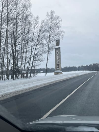 The image displays a winter roadside scene in Butyrki, Russia, viewed from inside a vehicle. A dark asphalt road, marked by a solid white center line, occupies the foreground and recedes into the distance, with snow partially covering its shoulders. To the right of the road, a prominent, tall grey monument stands on a snow-covered base. Its rectangular pillar bears vertical Cyrillic lettering that spells "БУТЫРКИ" (Butyrki), topped by a smaller, dark rectangular sign. To the left of the road, a dense cluster of bare deciduous trees, primarily birches, rises from snow-covered ground. Several small, snow-dusted structures are faintly visible through the trees on the left side of the frame. Beyond the monument and the road, a flat, snow-covered field extends to a distant treeline. The sky is uniformly overcast and grey. No individuals are present.