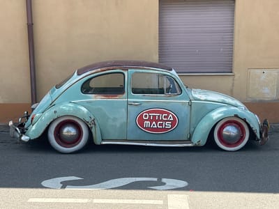 In Muravera, Italy, a vintage turquoise Volkswagen Beetle is parked on an asphalt street in front of a tan building. The car, showing signs of rust and wear, features white-walled tires with red rims. A red oval sticker with white lettering that reads "OTTTICA MACIS" is affixed to the driver's side door. The building has a closed, grey rolling shutter and a downspout running vertically on its left side. White road markings are visible on the street in the foreground.
