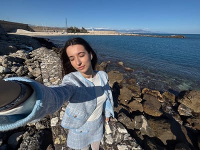 A young adult female is depicted in a selfie, standing on a rocky shoreline in Antibes, France, under a clear blue sky. She has long dark hair and a fair complexion, with her eyes closed and a faint smile. She is wearing a light blue tweed-style jacket with a matching mini-skirt, a white top, and white patterned tights. Her left arm is extended towards the camera, revealing a silver-colored bracelet on her wrist. A small white handbag is visible in her right hand. She stands on a cluster of large, dark brown rocks forming a breakwater, with the clear, blue-green Mediterranean Sea to her right. In the background, a curved stone seawall borders a sandy beach where several distant figures are visible. Beyond the beach, coastal structures and sailboat masts are discernible. In the far distance, snow-capped mountains are visible on the horizon. The scene is illuminated by strong sunlight, casting prominent shadows.
