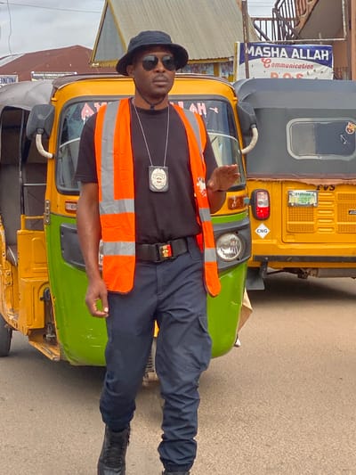 In Rigachikun, Nigeria, a Black man wearing a black bucket hat, dark sunglasses, a black t-shirt, an orange safety vest with reflective stripes, dark pants, and black boots stands on a street next to two auto-rickshaws. He has a badge hanging from a lanyard around his neck. One auto-rickshaw is yellow and green, and the other is gray. The man is gesturing with his right hand, palm facing outward. In the background, there are buildings and signs, including one that reads "MASHA ALLAH Communication POS." The street is paved with asphalt.