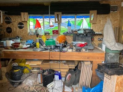 The image depicts a cluttered workshop in Walton-on-the-Naze, United Kingdom. A workbench dominates the scene, covered with tools, containers, and materials. Below the workbench is a vacuum cleaner, various pieces of wood, buckets, and tires. On the wall behind the workbench, circular saw blades are mounted, along with a stained-glass window depicting sailboats. Three wooden picture frames hang from the ceiling.