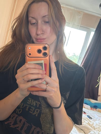 A young woman with auburn hair is taking a selfie in a room. She is holding an orange-cased smartphone in both hands, angled to capture her face and upper body. She is wearing a black t-shirt with text that appears to be a band logo. There are two rings on her fingers, and a nose ring. In the background, a window is visible with sheer curtains, and part of a bed is visible with white bedding. The lighting suggests it is daytime, possibly late afternoon or early evening. The setting appears to be a bedroom or living space. The image quality is moderate, with some noise and slight blur. The location in Ladozhskoe Ozero, Russia is not ascertainable from the visual information presented in the image.