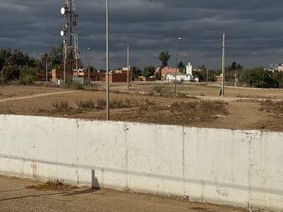 The image depicts a semi-urban landscape in Sidi Slimane, Morocco, under a heavily overcast sky. In the immediate foreground, a light grey concrete retaining wall runs horizontally across the lower third of the frame, showing vertical seams and some staining. Below the wall, a dark paved surface exhibits a prominent, elongated shadow cast to the right.

The midground consists of a large, undeveloped dirt field, characterized by dry, sparse, brown vegetation and visible dirt paths. Scattered across this field are several tall, slender grey street lamp poles. A prominent feature on the left side of the midground is a tall, metallic communications tower equipped with multiple white, dish-shaped antennas. To the right of the tower, a solitary figure, silhouetted in dark clothing, stands near a vertical pole.

The background reveals a mix of residential and natural elements. On the left, a cluster of green trees, including palm trees, partially obscures reddish-brown brick buildings, some appearing to be under construction. Towards the center-right background, a distinct white building with a green roof section and a small, white minaret-like structure is visible, surrounded by other less defined buildings and additional green foliage. The sky is dominated by dark, heavy grey clouds, indicating an overcast or potentially stormy condition.