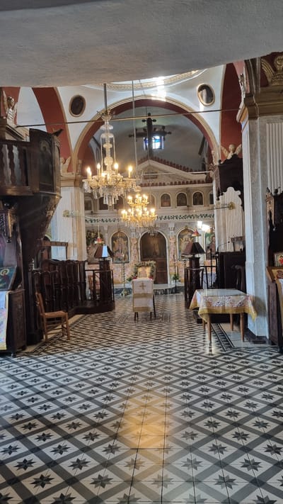The image shows the interior of a church in Palaios Panteleimonas, Greece. The floor is tiled with a geometric pattern of black and white diamonds and stars. In the foreground, a wooden chair with a woven seat is visible. Further into the church, there are two ornate chandeliers hanging from the ceiling. The walls are decorated with religious icons and paintings. In the center of the image, a white cloth-covered lectern with a cross on it stands. To the right, a small table is covered with a patterned tablecloth. The altar area, at the far end of the church, features more religious imagery. Sunlight streams in from an unseen source, casting a warm glow on the floor. The overall impression is one of a traditional and richly decorated place of worship.