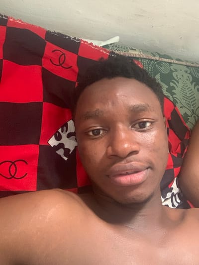 An indoor scene in Eziobodo, Nigeria, depicts a young adult male taking a selfie while lying down. The individual, of dark complexion with short dark hair, is positioned with his head resting on bedding. He looks directly at the camera with a neutral expression, his lips slightly parted. His bare upper chest and shoulders are visible. To the left side of the frame, a red and black checkered fabric with a black double-C logo resembling the Chanel brand is present. To the right, a green fabric with an intricate pattern is visible. A section of a light-colored wall is observable at the top of the frame. Another fabric featuring abstract white/grey and black shapes is visible near the subject's right shoulder.