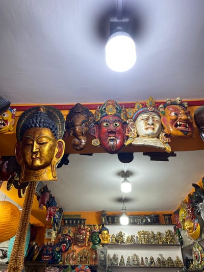 The image shows a display of Tibetan masks and statues for sale in a shop in Kathmandu Metropolitan City, Nepal. The masks are predominantly made of wood and are brightly painted in various colors and designs. Some masks depict faces with exaggerated expressions, while others have more serene features. The statues appear to be made of brass and represent deities and other figures from Tibetan Buddhism. The shop is dimly lit, with bright overhead lights illuminating the merchandise. Shelves are stacked with more items, including prayer flags and other religious artifacts. The overall impression is of a vibrant and culturally rich marketplace.