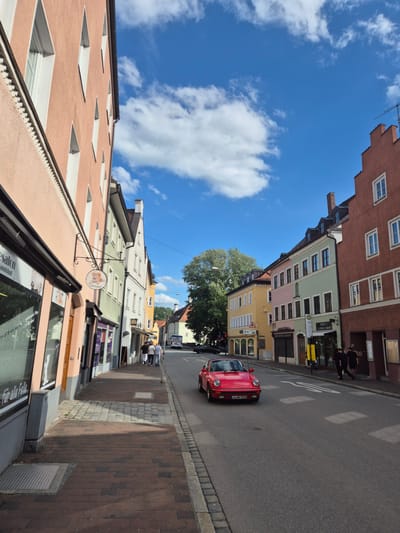 The image depicts a street scene in Landshut, Germany, specifically the Altstadt. The viewpoint is from a pedestrian's perspective on the left side of the street, looking down the street as it curves slightly to the right. The street is lined with colorful, historic buildings, characteristic of the Altstadt. The buildings are multi-story, with distinct architectural styles, featuring pitched roofs with dormer windows, arched doorways, and a variety of window shapes and colors. The facades are painted in pastel shades of peach, green, yellow, and pink, contrasted by a prominent reddish-brown building on the right side of the image.
The street is paved with asphalt, and there is a brick sidewalk on the left side, bordered by cobblestones. Various shops and businesses are visible on the ground floor of the buildings, indicated by signage and storefronts. One storefront on the left displays text that includes "für alle Felle," suggesting a pet-related business. Another sign, hanging from a bracket, features a circular graphic of a dog. Further down the street, a sign reads "Jimmy Barber Shop."
In the center of the street, a red Porsche car is driving towards the viewer. The car has its headlights on and a German license plate that reads "LA HH 911H". Several pedestrians are visible on the sidewalk. On the left sidewalk, three individuals are walking away from the camera. On the right sidewalk, two figures are walking towards the camera, and further down, a group of three people are walking away from the camera. A white bus is parked or driving on the right side of the street in the distance.
The sky is bright blue with scattered white clouds, indicating fair weather. A large tree with dense foliage is visible between the buildings on the left side of the street, further down. The overall impression is that of a charming, historic European city street on a sunny day.