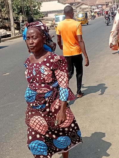 An elderly woman in a patterned dress and headwrap stands in the street, holding a small object. Behind her walks a younger man in an orange shirt and dark pants. A yellow vehicle is visible in the background, along with buildings and other people. The scene is lit by bright daylight, casting shadows on the pavement.