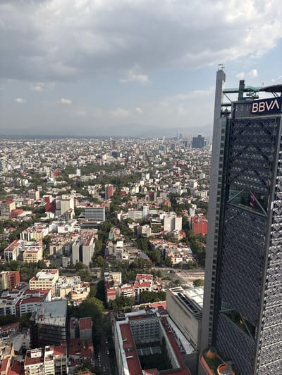 An aerial view captures a dense urban landscape under a cloudy sky, with a prominent modern skyscraper on the right side of the frame. The skyscraper, which appears to be the BBVA Bancomer Tower in Mexico City, is characterized by its dark, diamond-patterned facade and a prominent "BBVA" logo. The rest of the image showcases a sprawling city with numerous buildings of varying heights and architectural styles, interspersed with patches of green trees. Rooftops are predominantly red and white. The distant horizon is hazy, with mountains faintly visible. The city extends as far as the eye can see, suggesting a vast metropolitan area. There are no visible people or vehicles in the frame. The environment appears to be a bustling, developed urban center.