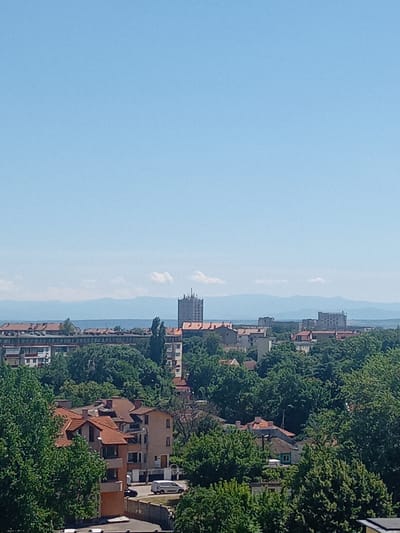 This is a high-angle, eye-level, medium shot of a cityscape, likely taken from an elevated position. The sky is a clear, bright blue, with a few wispy clouds visible in the distance. In the mid-ground, a sprawling urban area is densely packed with buildings of various sizes and architectural styles. Many buildings feature red-tiled roofs, characteristic of Vidin, Bulgaria. Numerous trees, lush and green, are interspersed throughout the urban landscape, creating a verdant atmosphere.
Dominating the skyline are several taller buildings. To the right of the frame, a prominent, light-colored, rectangular tower stands out, equipped with what appears to be communication antennas on its roof. Further to the left and in the background, other multi-story apartment buildings are visible, some with balconies. A long, low-rise building with a repeating window pattern runs horizontally across the mid-ground.
In the foreground, the roofs of several houses are visible, also with red tiles. A gray stone fence runs along the bottom edge of the image. Two vehicles are parked or driving on a road: a white van and a dark-colored car. The overall impression is of a moderately dense urban environment within a temperate climate, likely during the daytime on a clear day. The presence of red-tiled roofs and the mountainous horizon in the distance are consistent with the geographical features and typical architecture of Vidin, Bulgaria.