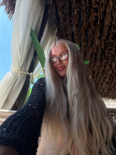 This media file shows a close-up, high-angle shot of a young woman lying down. She has long, blonde hair that frames her face, and she is wearing glasses with round, reflective lenses. Her eyes are partially closed, and her mouth is slightly open, suggesting a relaxed or resting state. She is wearing a dark, loosely knitted garment on her arm.
The background is composed of several elements. Above her, there is a thatched roof, possibly made of palm leaves, which creates a rustic texture. Below her, white curtains are draped, partially obscuring a view of a bright blue sky, indicating it is daytime. A green leaf is visible, emerging from behind the curtains, adding a touch of nature to the scene. The overall impression is one of leisure and relaxation, perhaps in a tropical or beachside setting. The scene is likely outdoors, given the clear sky and natural materials. No text is visible in the image. The location context provided is Drobnići, Montenegro, which aligns with a potential vacation or getaway scenario.