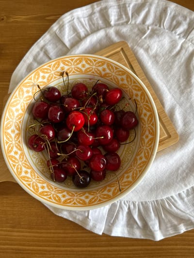 A top-down shot displays a bowl filled with dark red cherries. The bowl, featuring a yellow patterned rim, rests on a textured wooden surface. A white ruffled cloth is partially visible beneath the bowl and to its left. The lighting suggests it's daytime, with soft shadows indicating natural light. The scene is still and peaceful, focusing on the fresh fruit. There is no visible text.
