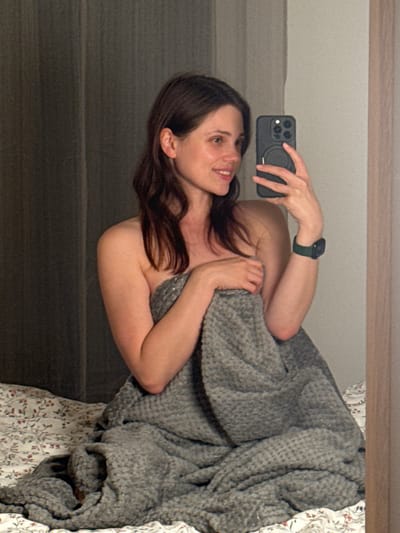 A woman is taking a selfie in a mirror. She is holding a black smartphone in her right hand. The woman is wearing a dark green smartwatch on her left wrist. She is partially covered by a gray textured blanket. The background includes a white bedsheet with a floral pattern and a gray wall. The location is not identifiable as Balatonlelle, Hungary, from the image alone.
