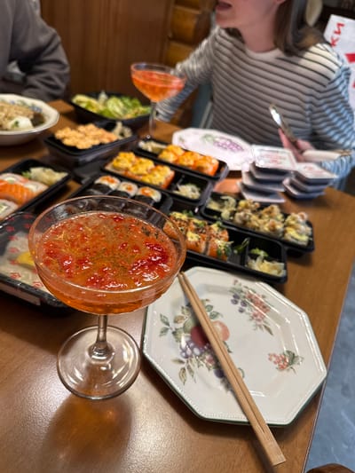 A person in a striped shirt is seen using chopsticks to pick up sushi from a variety of black plastic containers on a wooden table. A cocktail glass filled with an orange liquid is prominently displayed. A patterned octagonal plate with chopsticks on top is on the right side of the table. The location is Tomilino, Russia.