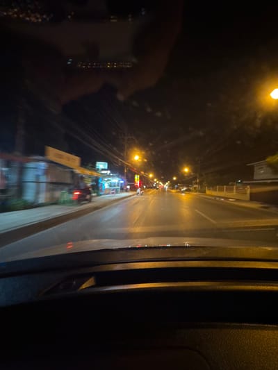 This is a nighttime, dashcam view of a street in Baan Lipa Noi, Thailand. The street is illuminated by streetlights and the lights from shops and buildings lining the road. On the left side of the road, there are small businesses with awnings. A dark-colored car is visible on the street, with its taillights illuminated. The road appears to be paved and wet, reflecting the light from the streetlights. There are power lines visible overhead. In the foreground, the dashboard of the vehicle is visible. The scene conveys a sense of movement, as indicated by the motion blur of the lights and buildings.
