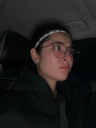 The scene is set inside a vehicle in Komchén, Mexico, characterized by dim lighting. A single young adult female is depicted, seated, facing slightly to the right of the camera. Her dark hair is contained by a white headband featuring a repeating pattern of black three-stripe logos. She wears circular, wire-frame spectacles and a dark-colored hooded sweatshirt. A black seatbelt is visible diagonally across her upper torso. A small, silver-toned, decorative earring is present on her visible left earlobe. Her eyes are directed upward and to the right, with a subtly furrowed brow and a closed mouth, indicating an expression of focus or mild apprehension. The immediate background shows a dark vehicle headrest and obscured dark interior components, consistent with a vehicle cabin.