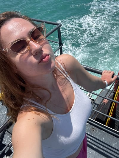 A woman with fair skin and red hair is taking a selfie from a ferry. She is wearing cat-eye sunglasses, a white tank top, and pink shorts. She has her lips pursed, as if kissing the camera. The water behind her is choppy and turquoise, with white foam from the ferry's wake. The railing of the ferry is visible in the foreground, as well as a part of the deck. The woman's right hand is resting on the railing. She is wearing a ring on her left hand and a white bracelet on her right wrist.