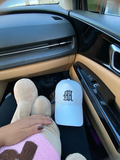 A woman's hand rests on a light brown stuffed toy. A white baseball cap with a black embroidered gothic "MH" logo is placed next to the toy. The scene is inside a car, with the dashboard, door panel, and window visible. The lighting suggests it is daytime. The woman's fingernails are painted a dark brown color.