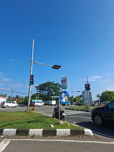 The image depicts a street intersection in Blahbatuh, Indonesia, under a clear blue sky. A traffic light pole with red and yellow lights illuminated is prominently featured in the foreground, along with a motorcycle parked on the side. Several vehicles, including cars and trucks, are visible on the road, some appearing to be stopped, possibly at the intersection. Green grass lines the roadside, and various signs and utility poles are scattered throughout the scene. The overall impression is of a busy urban environment on a sunny day.