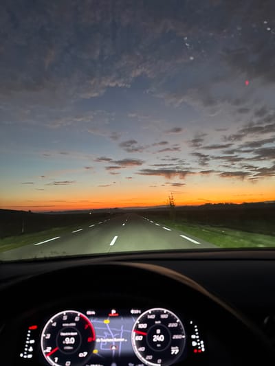 The photo depicts a car's dashboard and windshield view. The speedometer indicates a speed of 240 km/h, while the tachometer shows 9.0. The odometer reads 77. Through the windshield, a straight road stretches towards the horizon, flanked by green fields. The sky above is painted with hues of orange and pink, indicating either sunrise or sunset. Clouds are scattered across the sky, adding depth to the scenic view.