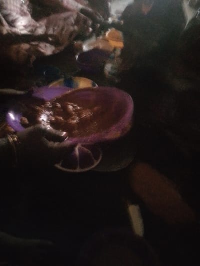 A purple bowl, likely containing a sauce, is held by a hand. The bowl is on a stand with a white rim. The image is dimly lit. A person is possibly seated near a couch in the background. The overall scene is unclear due to darkness and blur.