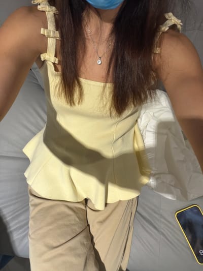 A woman is sitting indoors on a gray upholstered sofa. She is wearing a light yellow strapless top with tie details on the shoulders and tan trousers. A silver necklace with a small pendant is visible. A white cloth or fabric is beside her, along with a smartphone with a black screen and a yellow case. The location is Villahermosa, Mexico.
