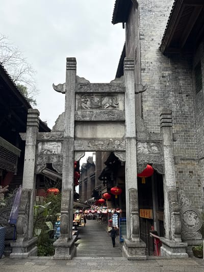 The image presents a stone archway (paifang) acting as an entrance to a historic street within Pengshui Miao and Tujia Autonomous County, China, under an overcast sky. The archway, constructed from weathered gray stone, features a central lintel with relief carvings of two stylized mythical creatures, possibly dragons, facing each other. Below this, smaller panels on either side of the archway display carved animal figures, potentially lions, amidst decorative patterns.
Beyond the archway, a stone-paved street, initially descending with steps, extends into the distance. It is flanked by multi-story buildings housing shops or stalls with traditional architectural elements. Numerous red spherical lanterns are suspended across the street overhead, continuing further down. A dense arrangement of colorful, tiered parasols or umbrellas also provides overhead cover further into the street. Several pedestrians are visible walking along the street, including one individual in a dark jacket walking away from the camera on the right side of the thoroughfare. A blue Pepsi branded refrigerator is visible on the right side of the street, just past the archway.
To the immediate right of the archway, a multi-story building constructed of gray brick with dark wooden eaves rises prominently. To the left, a darker building with traditional Chinese rooflines and visible dark wooden structural elements is partially observable. Bare tree branches are silhouetted against the gray sky in the upper-left quadrant, and green foliage grows near the base of the archway on the left side.