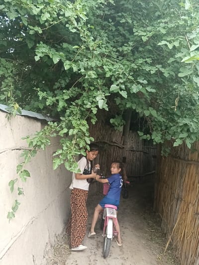The image depicts two young girls outdoors, positioned near a wall and a thatched structure. The girl on the left, wearing a white t-shirt and patterned pants, stands and appears to be interacting with the girl on the right. The girl on the right is seated on a pink bicycle, wearing a blue t-shirt and blue shorts, and is looking towards the camera. Lush green foliage hangs overhead, suggesting a shaded outdoor space. The background reveals a wall constructed from what appears to be mud or plaster, and a structure made of reeds or straw. The ground is dirt. The scene is set in the city of Altinkul, Uzbekistan.