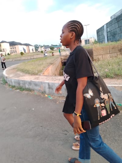 A young woman is walking on a paved road in Umueshi, Nigeria. She has her hair braided and is wearing a black short-sleeved shirt and blue jeans. She carries a black tote bag with cartoonish figures printed on it over her shoulder and a phone in her hand. Other people are visible in the background near a two-story building. The environment includes a concrete barrier, grassy areas, and a drain ditch. The sky is overcast.
