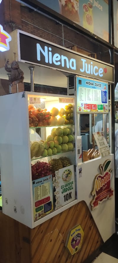 A street food stand at the coordinates -7.467, 112.438 in Mojokerto, Indonesia, displays a variety of fruit, including green apples, dragonfruit, and pineapples. The sign reads "Niena Juice," and a price list for various drinks is displayed. In the background, a person is visible, partially obscured by the stand. The stand features a white wooden frame, with a sign above it that reads "Niena Juice." Below the sign is a wooden panel with a price tag displaying the cost of a drink. The stand is located in a busy city center. The stand is lit by overhead fluorescent lights. The stand is also decorated with a few decorative items, including a small straw bird perched on the sign.