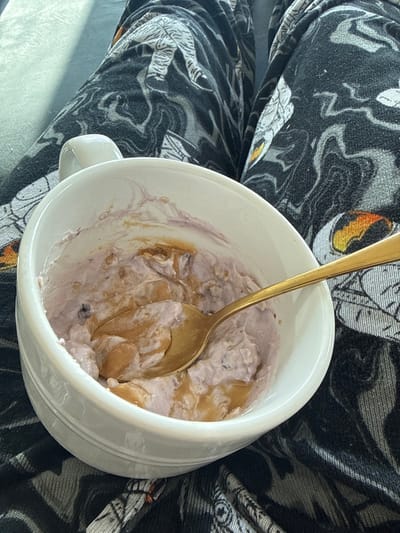 An overhead shot captures a white mug filled with a partially eaten meal, resting on a person's lap. The mug contains a creamy, light purple-hued substance, possibly yogurt or overnight oats, which is swirled with generous dollops of a lighter brown, thick consistency, indicative of peanut butter or a similar nut butter. Darker specks are visible within the purple base, suggesting berries or other mix-ins. A long, slender golden spoon is plunged into the mixture, angled towards the left, with some of the food clinging to its bowl.

The mug has a handle visible on its left side. Surrounding the mug, and serving as the background, is a person's leg or lap, covered by a piece of fabric. This fabric features a dynamic pattern of swirling black and grey, interspersed with white organic shapes resembling leaves or abstract motifs, and occasional subtle hints of orange. The lighting appears natural, casting a soft shadow on the bottom left corner of the fabric. There is no visible text in the image. The scene suggests a casual meal being consumed indoors.