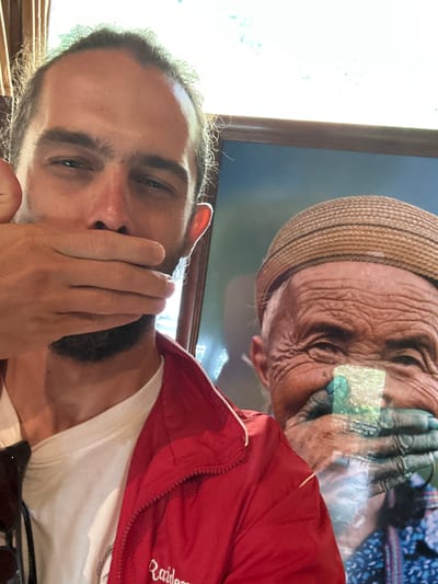An individual is depicted in a selfie, posing in front of a framed portrait in Púng Luông, Vietnam. The foreground shows a light-skinned male with a beard and dark hair, wearing a white t-shirt and a red track jacket with "Raiders" partially visible on the chest. His left hand is raised, covering his mouth and lower nose, mirroring the subject in the background. His eyes are open, directed towards the camera. Behind him, a framed picture displays an elderly, heavily wrinkled individual wearing a light brown woven hat with dark stripes. This person also has their hands covering their mouth and displays a smile. A distinctive rectangular patch of light green and white pigment is visible on the subject's lower right cheek in the portrait. The background of the portrait is a soft blue, and the frame is dark brown. The lighting suggests an indoor setting with bright ambient light from behind the subject on the left.