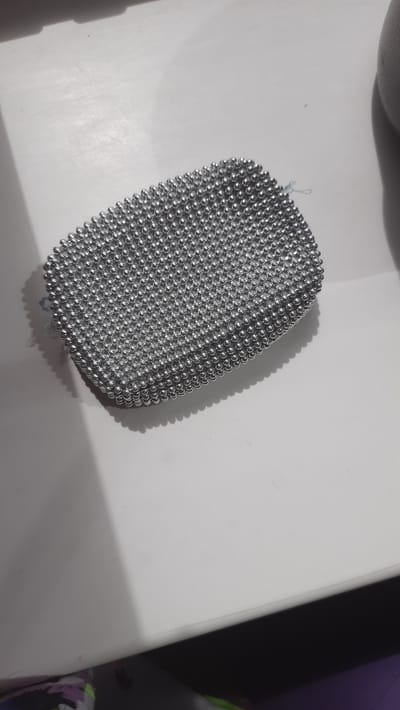 A close-up, high-angle shot shows a silver, beaded clutch bag resting on a white surface. The bag is oblong in shape with a slight curve and is covered in hundreds of small, spherical silver beads that create a textured, mosaic-like appearance. The beads are densely packed, forming a shimmering surface. The bag is positioned slightly off-center to the left of the frame, casting a soft shadow to its right. The white surface appears to be a table or countertop. The background is out of focus, but suggests a room with some ambient light and darker areas, possibly indicating furniture or other objects. The overall scene is clean and well-lit, with the focus sharply on the beaded bag. There is no indication of a specific location in Desnogorsk, Russia, or any interaction or action occurring.