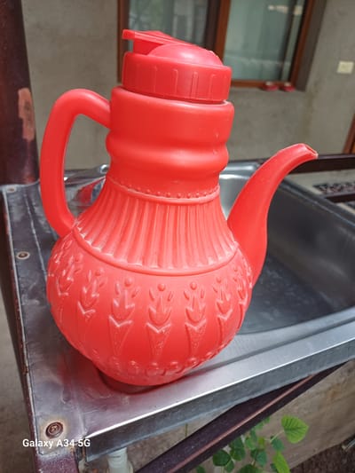 A bright red plastic teapot sits on a metal surface, likely a sink. The teapot has a decorative pattern of vertical lines on its upper body and a design resembling stylized leaves or flames on its lower body. A matching red lid with a small handle is securely fastened to the top. The spout of the teapot is curved and angled upwards. The metal surface appears worn and slightly stained, with visible rivets and edges. In the background, a blurred window with a dark frame is visible, and some greenery can be seen at the bottom right. The scene is outdoors, suggesting a domestic or industrial setting where such an object might be used or stored. The "Galaxy A34 5G" watermark at the bottom left indicates the image was captured with a Samsung smartphone. The location is identified as Angren, Uzbekistan.
