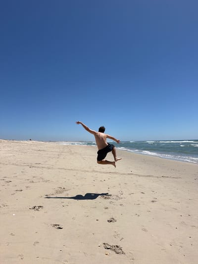 A person is captured mid-air, jumping on a sandy beach under a clear blue sky. The individual, wearing dark shorts, is shirtless, and their body is angled away from the camera. Their right arm is extended outwards and upwards, while their left arm is bent towards their body. Their legs are bent at the knees, with one leg slightly in front of the other, suggesting movement. The sandy beach stretches out towards a calm ocean with gentle waves. In the distance, the coastline is visible, with a few scattered individuals and structures. The foreground of the beach shows numerous footprints in the sand. The overall scene depicts a moment of freedom and exhilaration in a natural coastal environment.