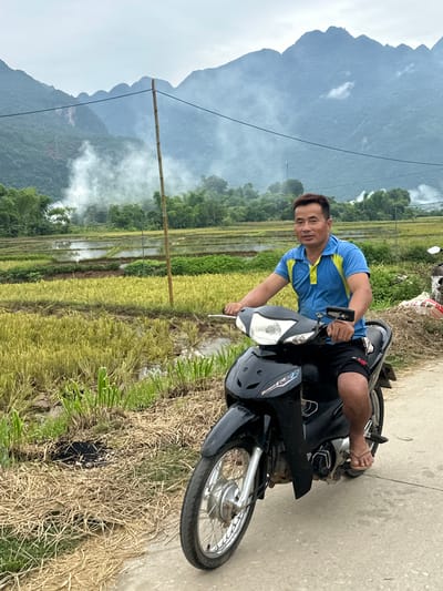 A middle-aged East Asian man, wearing a blue and yellow short-sleeved polo shirt and black shorts, rides a black motorcycle on a paved road. He is looking forward, with a neutral expression. The motorcycle has a silver front wheel and a black body. The man is wearing flip-flops.
The background shows a rural landscape in Mai Chau, Vietnam. There are rice paddies, some with harvested crops, and some with standing water. Green trees line the horizon. In the distance, mist partially obscures lush, green mountains. A tall, thin bamboo pole stands vertically to the left of the motorcycle. There is a visible power line stretching across the upper part of the image. The sky is overcast.