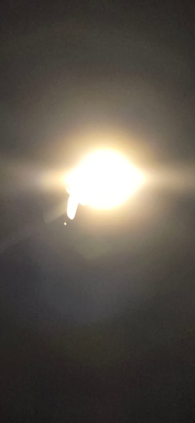 The image is a close-up of a bright light source, possibly a light bulb or a lamp, against a dark background. The light is very intense, appearing as a brilliant white-yellow glow that dominates the center of the frame. There is a lens flare effect, with streaks of light emanating from the source and scattering across the image. To the lower right of the main light, there is a smaller, distinct point of light, which could be another light source or a reflection. The background is dark and appears to be a solid color, with some subtle gradations in tone, suggesting it might be a wall or a ceiling. The overall impression is of a very bright light in an otherwise dark environment. There are no discernible subjects, activities, or explicit location cues within the image itself, other than the possibility of an indoor setting due to the nature of the light source. The lighting suggests it could be nighttime or a dimly lit interior space.