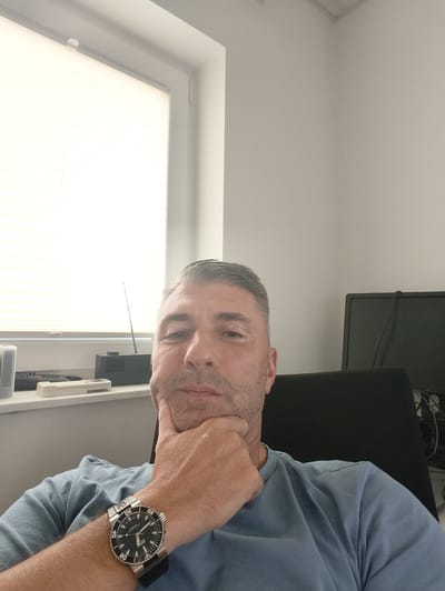 A man with short, graying hair and stubble is looking at the camera with his right hand resting on his chin. He is wearing a blue t-shirt and a silver watch with a black band. The man is sitting in an office chair in front of a desk. To the left of the man is a window with white blinds. On the desk, there is a black radio, a black computer monitor, and other office supplies. The environment appears to be an office in Mannheim, Germany.