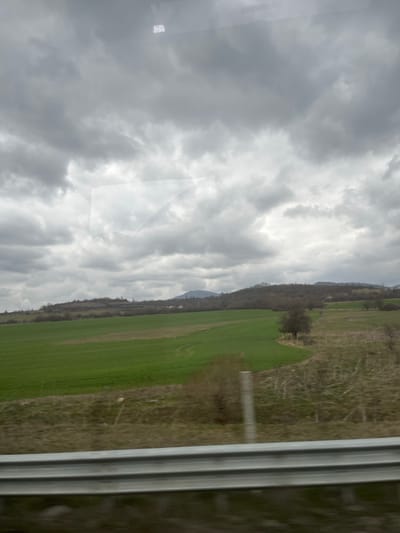 The image depicts an overcast rural landscape in Dzhurovo, Bulgaria, observed from a moving vehicle. The scene is dominated by a heavily clouded, grey sky, occupying approximately the upper two-thirds of the frame. Below the sky, a panoramic view of agricultural fields and rolling hills extends across the midground. The central and left foreground features a vibrant green field, indicative of a cultivated crop, with subtle ground patterns visible. To the right, the terrain transitions to drier, brownish-green scrubland with sparse, deciduous bushes and a single, isolated, bare-branched tree. In the mid-distance, a line of hills, largely covered with bare trees and some patches of dormant vegetation, runs horizontally, with faint structures resembling distant houses or buildings nestled on their slopes. Further in the background, a distinct, darker mountain peak is visible beneath the heavy cloud cover. The extreme bottom foreground shows a blurred, metallic guardrail, indicating the viewpoint from a roadside. No people, animals, or active interactions are present in the scene.