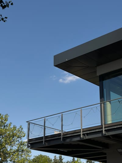 The image is a low-angle shot looking up at modern architecture against a clear blue sky. The primary subjects are the angular, dark-colored facade of a building and a metal safety railing with a wire mesh, illuminated by sunlight. The building features clean lines and dark, rectangular panels, with a prominent overhang. The railing appears to be part of a balcony or walkway, constructed from metal supports and a dense wire mesh, held taut by metal cables.
The scene is outdoors on a bright, sunny day, indicated by the clear sky and the way the sunlight highlights the architectural elements. There are no people visible in the image. The setting appears to be urban, given the modern construction, and the presence of lush green trees at the bottom right suggests a location with some natural elements, possibly a park or a landscaped area near the building. A few wisps of white cloud are visible in the sky.
There is no discernible activity taking place; the image captures a static architectural view. The overall mood is one of clean, modern design and a bright, pleasant day. The location is specified as Frankfurt, Germany, which aligns with the contemporary architectural style. No text is visible in the image.