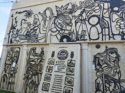 This image captures a large public monument situated in Dura, Palestinian Territory, under a clear daytime sky. The monument consists of a light-colored stone wall extensively decorated with black metal cutouts, forming a multi-panel mural.
The upper section presents a continuous frieze with stylized, flat-profile figures and natural elements. On the left, a sun, multiple birds in flight, a horse, a robed figure with a head covering, and a large cluster of grapes are visible. Adjacent to these are prominent figures, including one with flowing hair. The right portion of the upper frieze integrates geometric shapes, grapevines, a cluster of grapes, and a stylized human figure holding an object aloft. The word "DURA" is incorporated into the design in the upper right quadrant.
The lower section of the monument is composed of several distinct vertical panels, separated by vertical stone pillars. The central panel, positioned at a slight angle, features a circular portrait at its apex. Below the portrait, Arabic text is displayed alongside a series of square vignettes depicting symbolic images such as a sunrise over water, leaves, grapes, and an open book. A white cylindrical lantern is suspended in front of this panel. The panel to the left of the central one exhibits abstract black metal cutouts, with another white cylindrical lantern hanging in front. The panel to the right of the central one portrays a large, stylized female face or head adorned with a traditional scarf, accompanied by a dove and elements resembling wheat stalks.
The monument is erected on a patch of green grass at its base. Utility wires are visible across the top and along the sides of the structure.