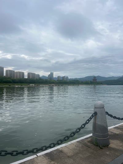 The image depicts a waterfront scene in Liandu District, China. In the foreground, a concrete pathway lines the edge of a body of water. A grey granite bollard with horizontal grooves and a rounded top is situated on the pathway, connected to a heavy black metal chain that extends along the water's edge. The water is calm, with ripples creating a textured surface that reflects the overcast sky.
Across the water, the opposite bank features a line of modern apartment buildings with multiple stories. Behind the buildings, a lush green treeline separates the urban development from rolling hills. Further in the distance, a multi-tiered pagoda-style building with a dark, ornate roof stands prominently against the backdrop of the hills. The sky is heavily clouded, contributing to a muted, diffused light. No people are visible in the image.