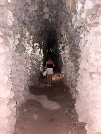 The image captures a top-down view of a person inside a deep, narrow shaft or cave. The walls of the shaft are extensively covered in light-colored, rough, and irregular formations, resembling crystallized deposits or small, knobby stalactites, creating a highly textured environment. The lighting is dim, suggesting an enclosed subterranean space or nighttime, with light appearing to come from the opening above or an artificial source near the subject.
A woman is visible lying on her back near the bottom of the shaft, looking upwards towards the camera. She has dark hair and is dressed in a black top or swimsuit and white shorts, with light-colored shoes. A white helmet with dark accents is positioned near her head or chest. Her posture suggests she is either resting, in the process of descending or ascending, or exploring the depths of the shaft.
The visible floor beneath the woman appears to be dark earth or rock. The overall scene conveys a sense of exploration within a unique geological feature or a man-made subterranean structure. Given the context of Cam Ranh, Vietnam, this could be a cave system, a deep well, or an old tunnel known for its distinct mineral formations. No text is visible in the image.