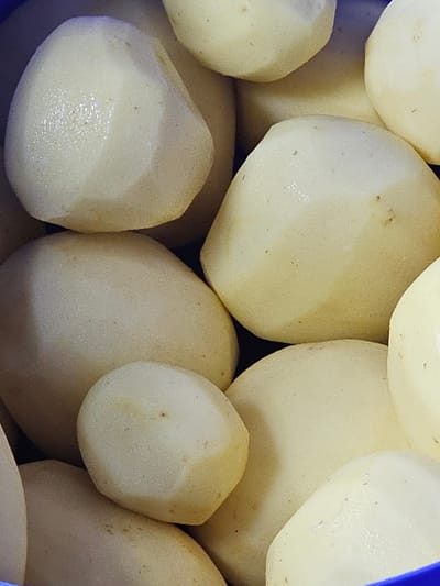 The image shows a close-up of a collection of peeled potatoes. The potatoes are various sizes and shapes, with a predominantly pale yellow to cream color. Some of the potatoes have small blemishes or marks. The background is dark and out of focus. No people or specific objects are visible to suggest a location in Holywell, United Kingdom.