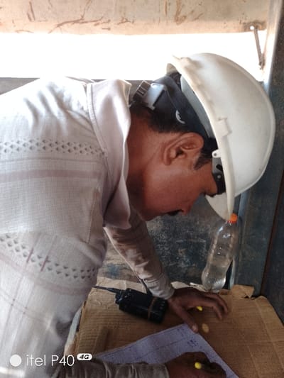 An individual is depicted indoors at Hoche, Nigeria, bending forward over a surface. The person wears a light-colored, long-sleeved collared shirt with patterned horizontal stripes across the chest and geometric designs on the sleeves, and a white hard hat with internal dark grey suspension straps. The hard hat is tilted upward on the head, revealing dark hair and the right ear. A dark-colored portable radio with an antenna is positioned on a brown, corrugated cardboard surface to the left of the person's right hand. The person's right hand, with visible knuckles, rests near two small, yellow, oblong objects on the cardboard. The left hand holds a yellow-capped pen and is actively writing on a light blue or white sheet of paper that displays visible text and lines. A partially consumed clear plastic water bottle with an orange cap is suspended vertically from the hard hat, positioned between the hard hat's brim and the person's right cheek. The background consists of a dark, possibly concrete or painted wall behind the person, and a lighter, distressed upper wall section. The floor beneath the cardboard appears dark and possibly dirty. Natural light enters from an unseen source, illuminating the scene.