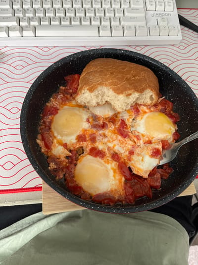 An indoor scene in Zainsk, Russia, displays a black cooking pan, speckled with white, containing four fried eggs nestled in a vibrant red tomato-based sauce with visible pieces of red pepper or sausage. A substantial, light brown piece of bread, with a distinct bite mark on its side, rests on top of the food. A silver spoon is partially submerged on the right side of the pan. The pan sits on a light-colored wooden trivet, which is placed on a white desk mat featuring a repeating red wave pattern. In the background, a white mechanical keyboard with Russian Cyrillic and Latin characters is visible, bearing the "ARDOR" brand logo in its upper right corner. Below the pan, a light green fabric, consistent with clothing, is present. No individuals are explicitly visible, but the arrangement suggests a person is consuming the meal at a desk or table.