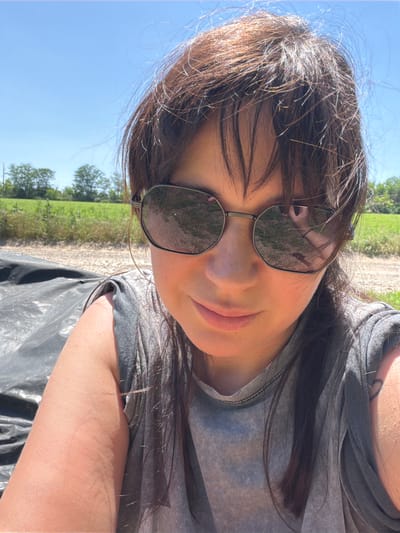 A selfie taken outdoors on a sunny day shows a woman wearing sunglasses and a grey t-shirt. She is lying down, with a black tarp and a field of grass visible in the background. The reflection in her sunglasses shows she is holding a phone and taking the picture. The woman has dark brown hair and fair skin.